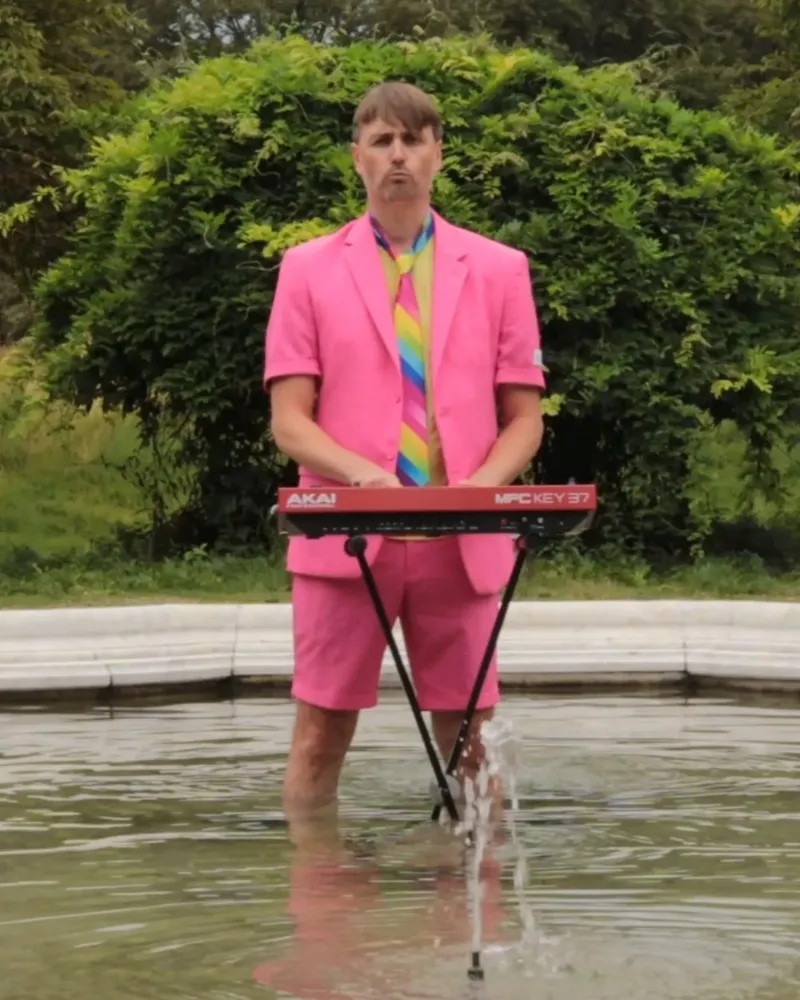 Andy Sauerwein im pinken Anzug mit Regenbogen-Krawatte steht mit einem Keyboard in einem Brunnen